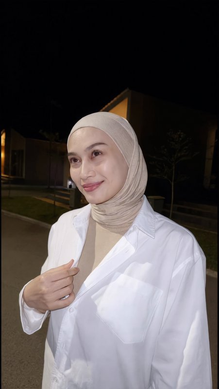 Smiling Muslim woman in a beige hijab and white button-up shirt poses for a casual outdoor night ...
