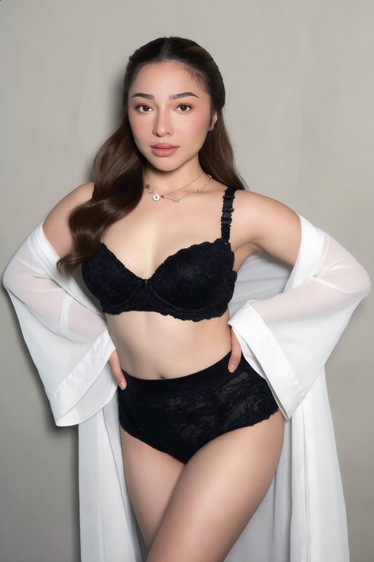Woman wearing black lace lingerie and sheer white robe poses against a plain gray studio wall