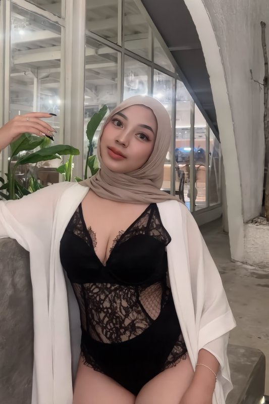 Woman in a beige hijab, black lace bodysuit and sheer white cover-up poses in a bright modern indoor lounge