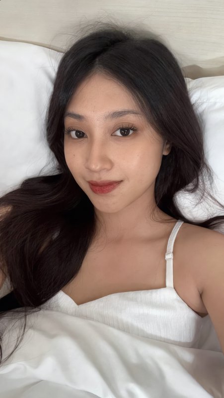 Young woman with long dark hair takes a casual selfie while lying against white bed pillows