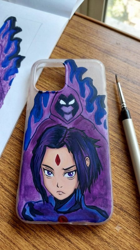 Hand-painted clear phone case with custom Teen Titans Raven fan art rests on a wooden desk next t...