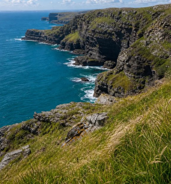 [High angle view of rugged grassy coastal cliffs plunging into bright turquoise Atlantic ocean on a sunny partly cloudy day]