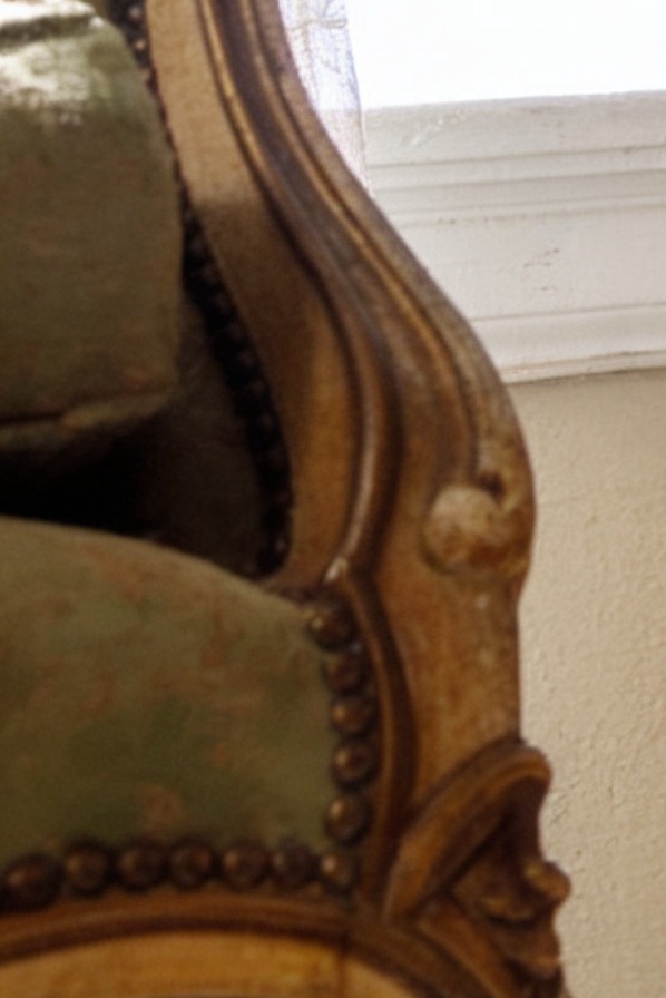 [Close-up of ornate carved gilded vintage armchair frame with sage green upholstery and brass nailhead trim, near a bright...