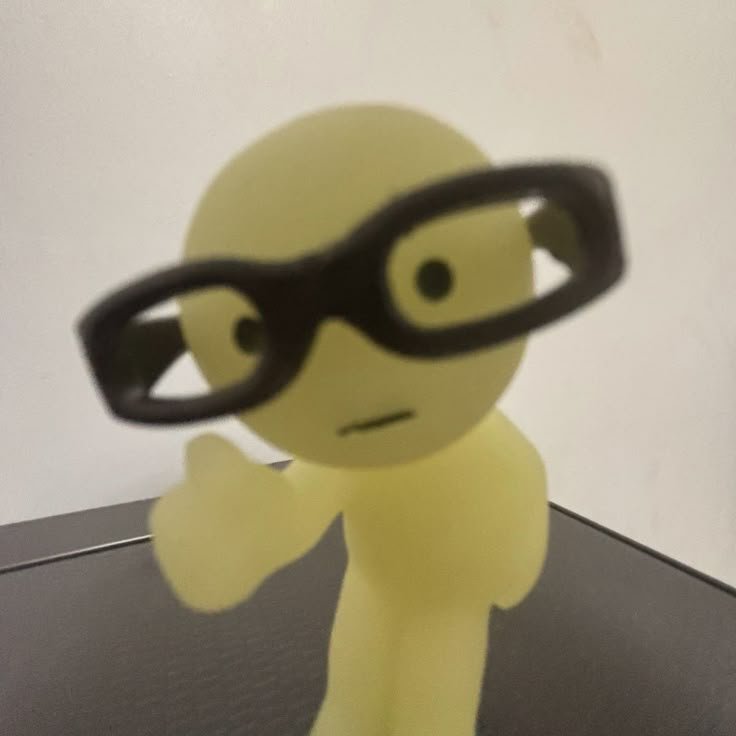 Blurry close-up of pale yellow humanoid blob figurine wearing large black glasses, giving a thumb...