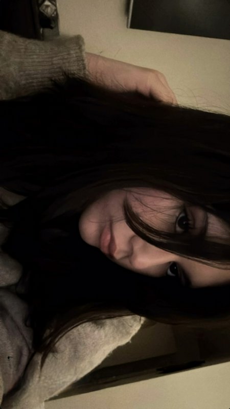 Low-light casual indoor selfie of a young woman with long dark hair, wearing a cozy knit sweater,...