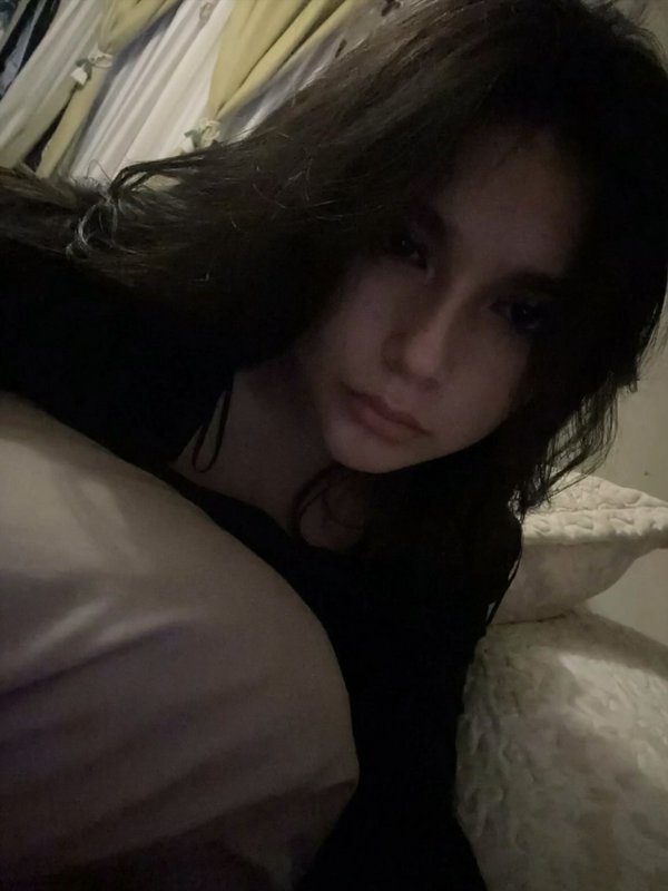 Dimly lit casual selfie of a young woman with messy dark hair resting in bed, looking toward the ...