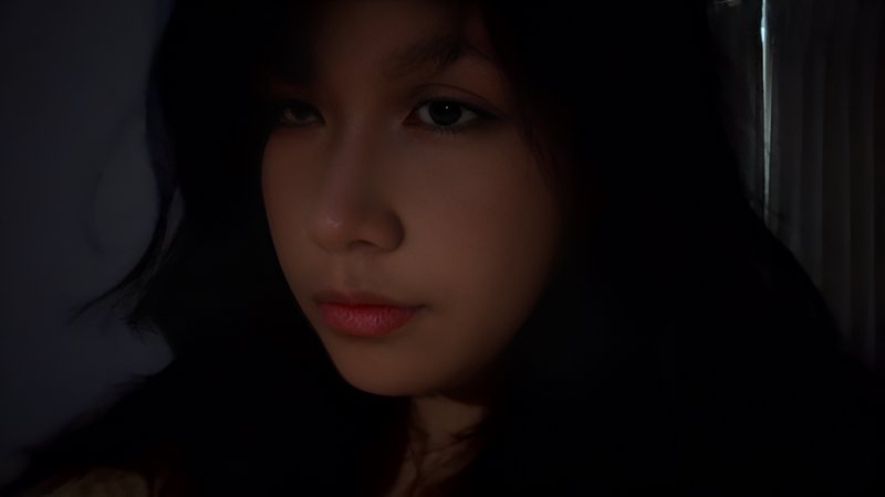 Close-up low-light portrait of a pensive young woman with dark hair, lit by soft warm light against a dark indoor background.
