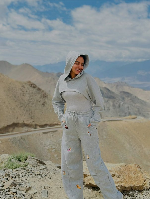 Smiling woman in grey embroidered hooded loungewear stands at a rocky high altitude mountain over...