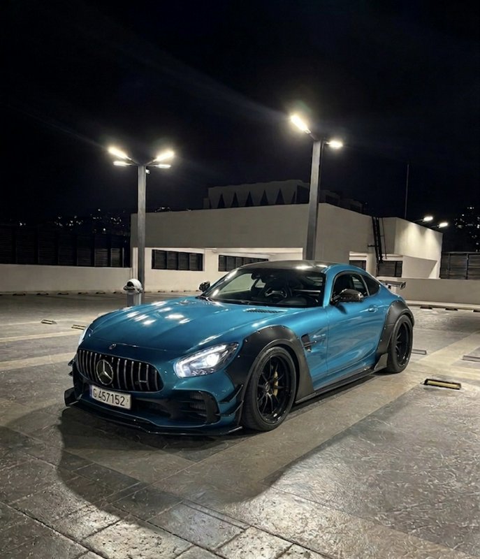 Widebody teal Mercedes AMG GT sports car parked on a paved rooftop parking deck at night under gl...