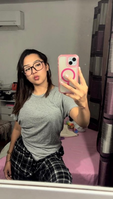 Young woman with oversized glasses takes playful bedroom mirror selfie holding a pink cased iPhon...