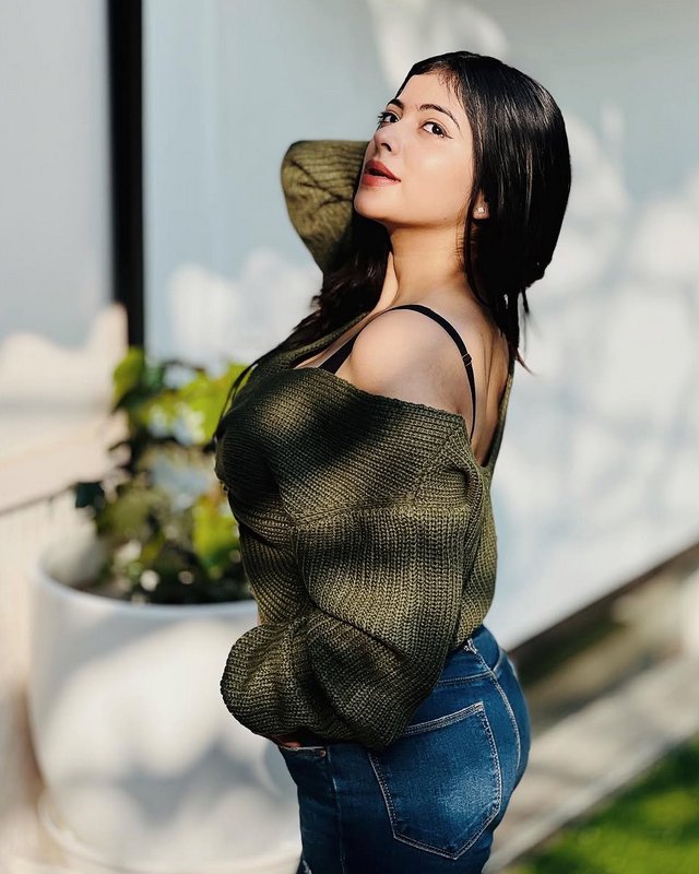 Young woman with sleek dark hair poses in warm sunlit patio wearing an off-shoulder olive knit sw...