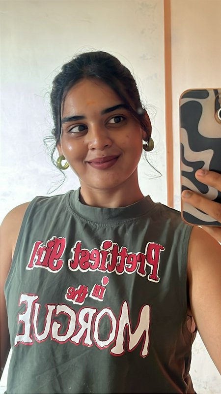 Smiling young woman takes mirror selfie wearing olive graphic tank top, green hoop earrings, hold...
