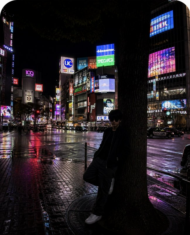 Silhouetted person leaning on a tree at night on rain-soaked Shibuya Crossing, Tokyo, with glowin...