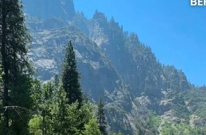 [Rugged granite mountain peaks tower over lush evergreen forest under a bright, cloudless sunny blue sky]