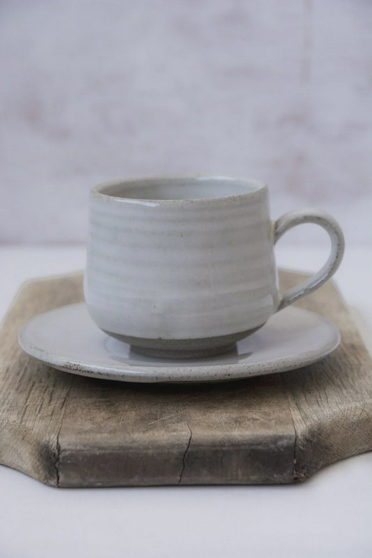 Handthrown ridged white ceramic coffee cup and matching saucer resting on a weathered rustic wood...