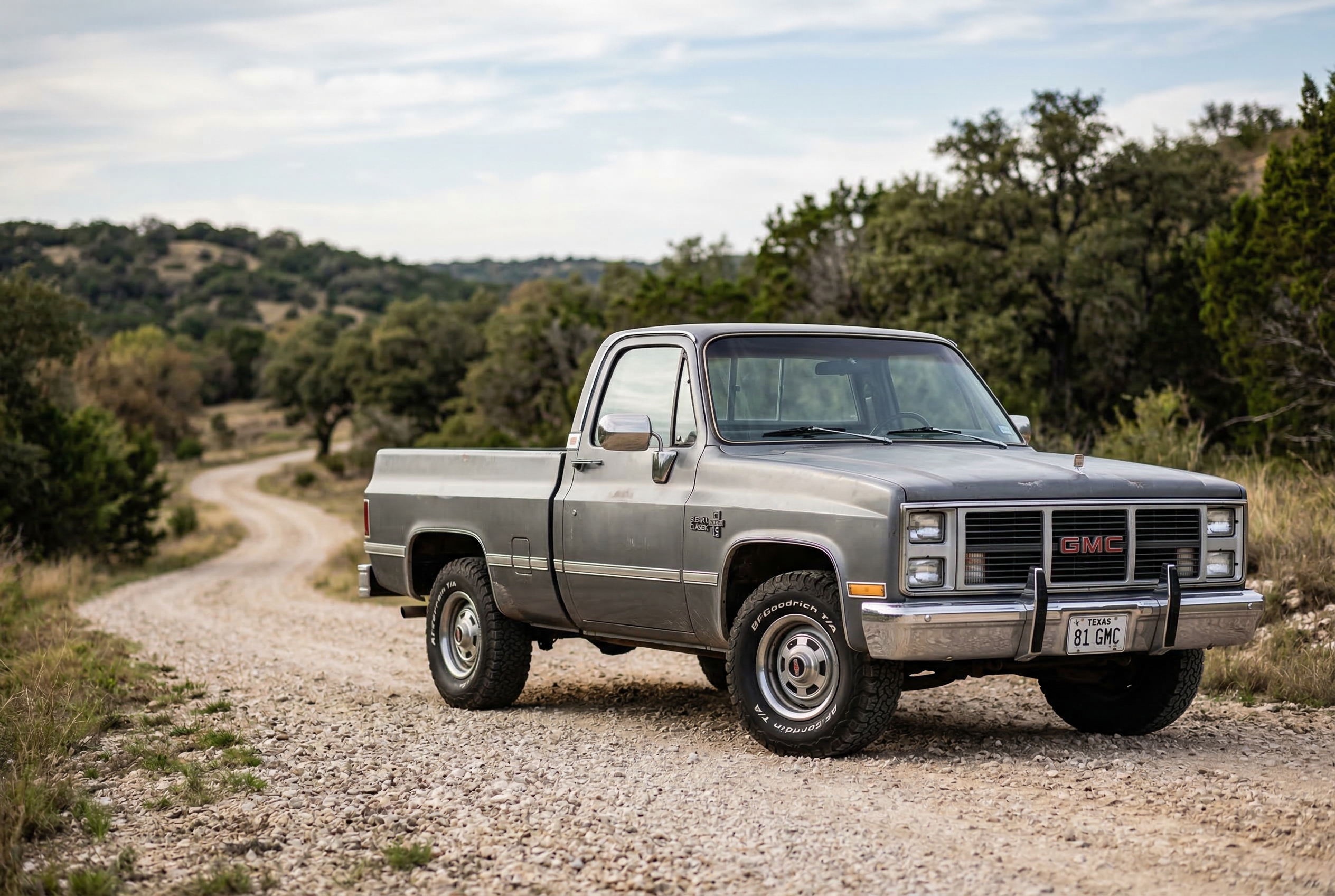 Retro gray GMC pickup parked on a gravel country road with hills and trees.