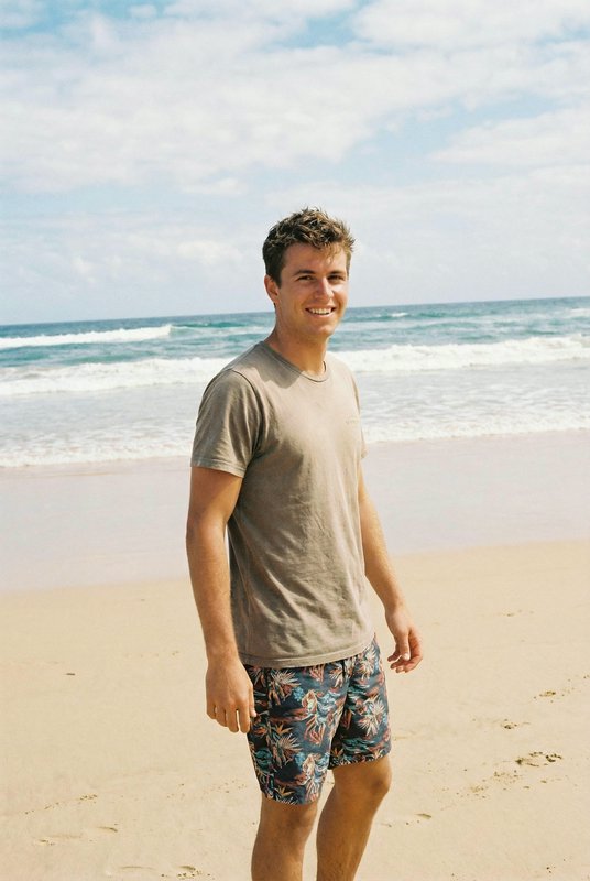 Man stands on sunny beach in beige t-shirt and tropical print swim trunks, ocean waves behind