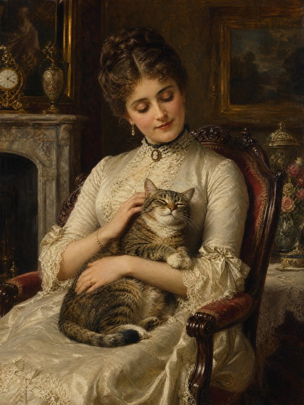 Woman in a lace ivory gown sits in an ornate chair, gently holding a content tabby cat.