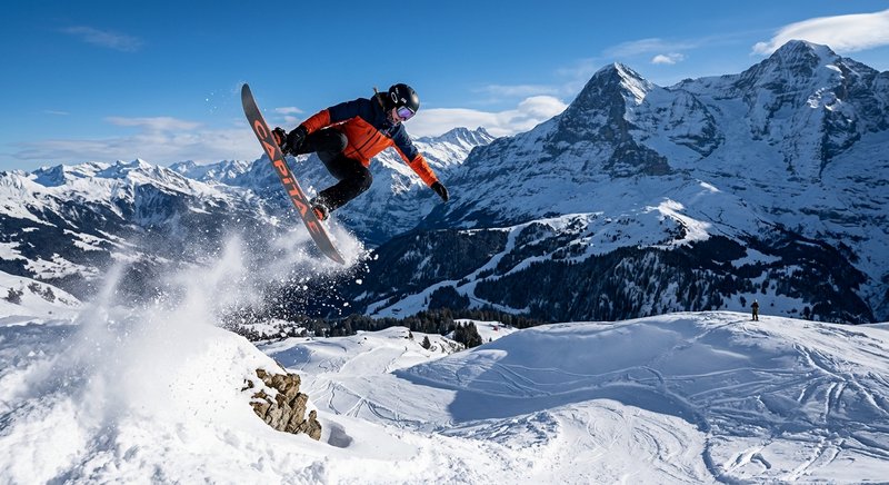 Snowboarder in orange jacket leaps off a snowy ledge with alpine mountains behind.