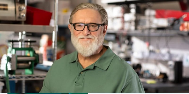 Senior bearded man with black glasses in a green polo, standing in a blurred industrial workshop ...