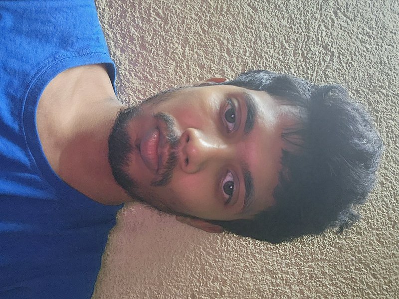 Close-up selfie of a young bearded South Asian man in a blue t-shirt, posed against a textured be...