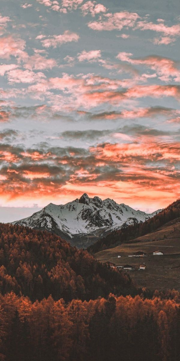 Snow-capped mountains rise above a fiery sunset over a forested valley.