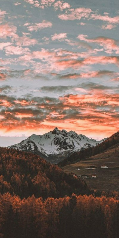 Snow-capped mountains rise above a fiery sunset over a forested valley.