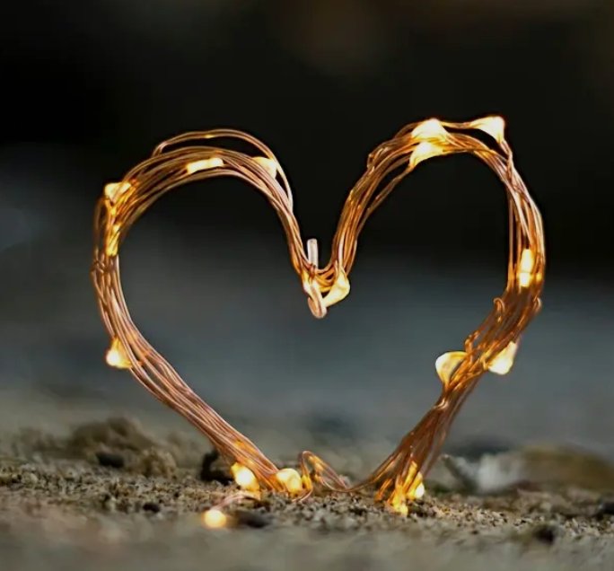 Wire heart shaped with glowing string lights on sand at night.