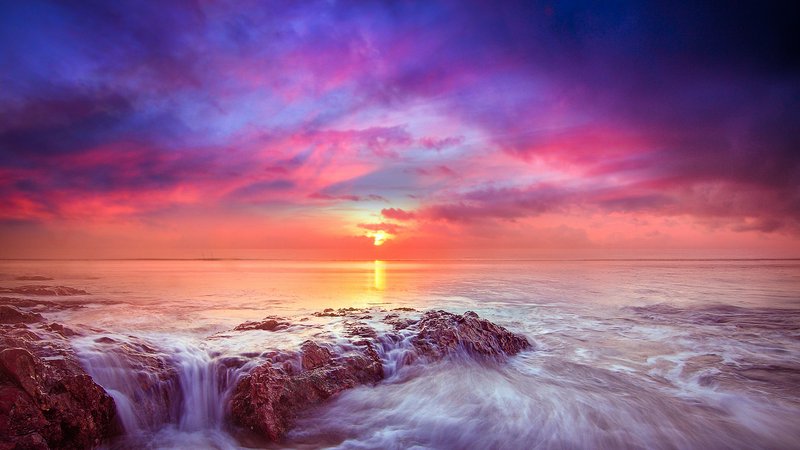 Dramatic vibrant ocean sunset, streaked purple and pink clouds above silky flowing waves washing over a rocky shoreline