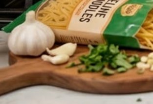 Garlic bulbs, parsley, and shredded pasta on a wooden cutting board with a pasta bag in the backg...