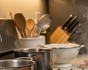 Stainless pots simmer on a stove with steaming steam and assorted wooden and metal kitchen utensi...