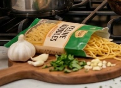 Bag of fusilli pasta spilling from a green-labeled pack onto a cutting board with garlic and pars...