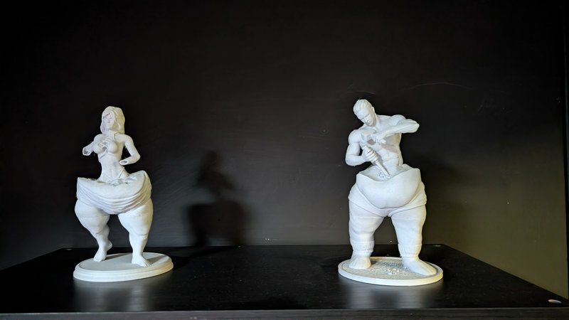 Two surreal white conceptual figurine sculptures displayed on a glossy black shelf against a plai...