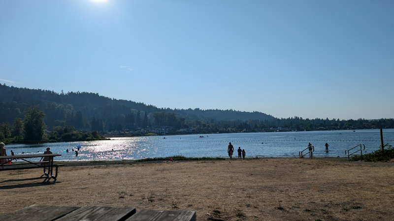 Bright sunny summer day at a public lake beach, visitors swim and relax on a sandy shore backed b...