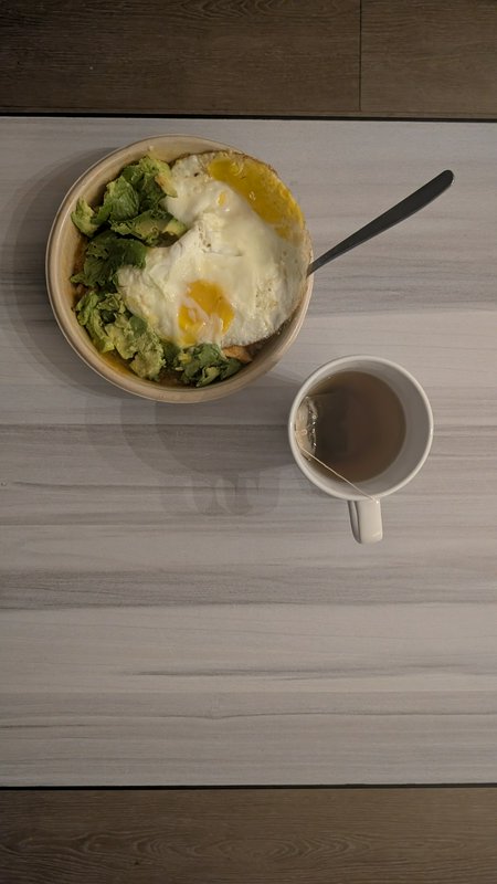 Overhead flat lay view of a breakfast bowl with fried egg, avocado, and a mug of hot tea on a lig...