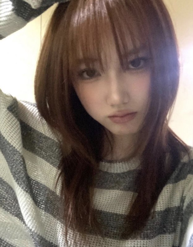 Close-up candid selfie of a young woman with light brown wispy bangs wearing a grey and white str...