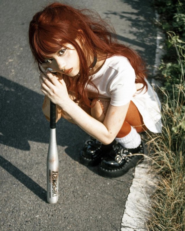 Red haired alt girl crouching on a roadside, holding a silver baseball bat and looking up at the ...
