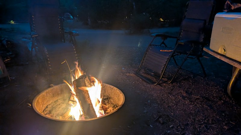 Flickering campfire in a metal fire ring at twilight, surrounded by empty folding camping chairs ...