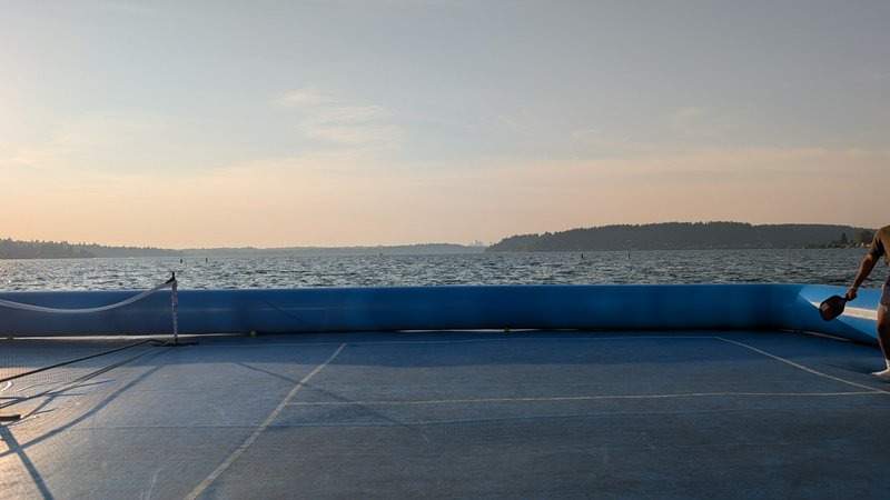 Inflatable floating pickleball court on a calm lake at golden sunset, partial player holding a pa...