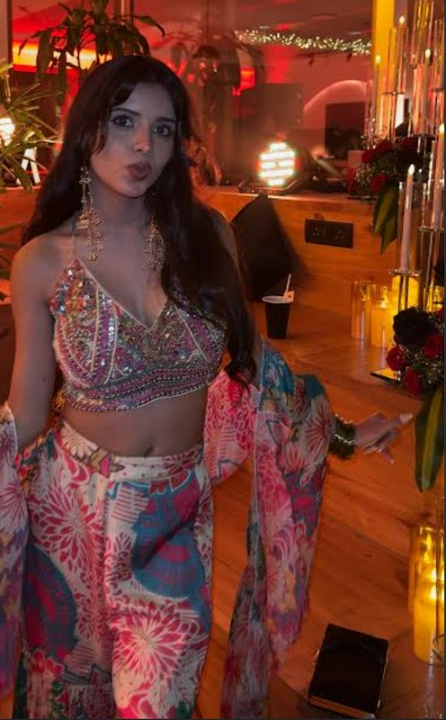 Young woman in embellished fusion ethnic party wear poses playfully at a warm red-lit indoor cele...