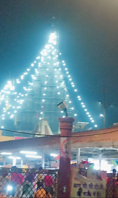 Illuminated Hindu temple spire strung with fairy lights rises above a crowded pilgrimage site at ...