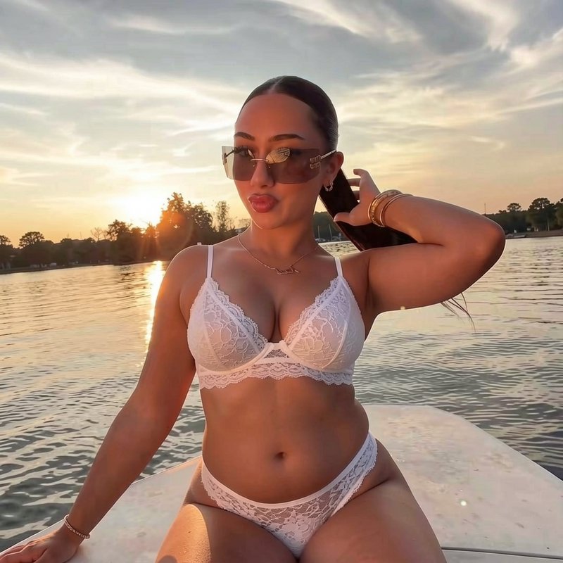 Woman in white lace bikini sits on boat bow as warm golden sunset glows over calm lake waters beh...