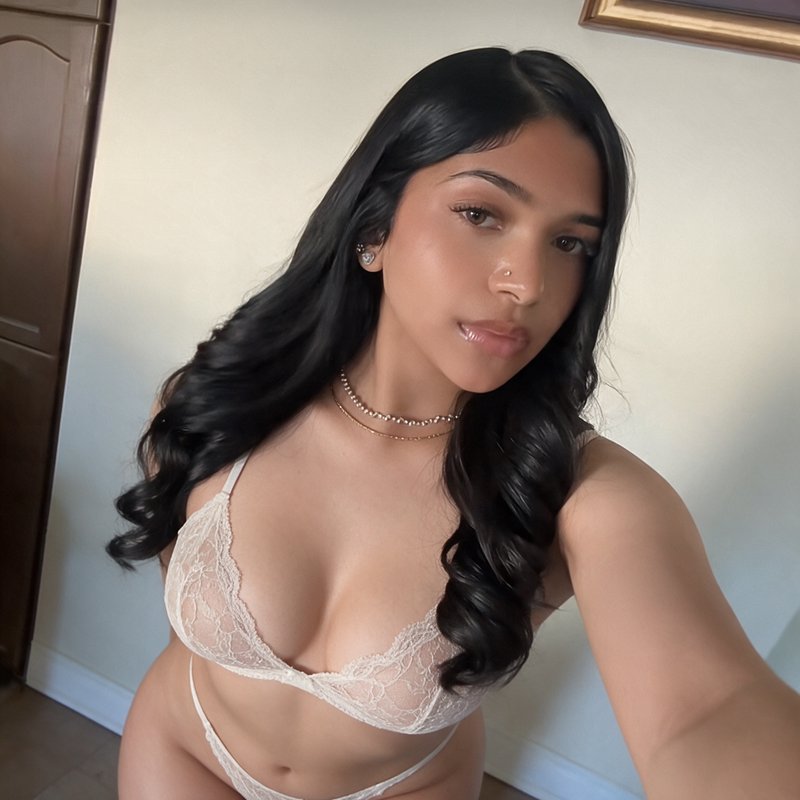Young woman with long wavy black hair takes a selfie wearing delicate white lace lingerie in a su...