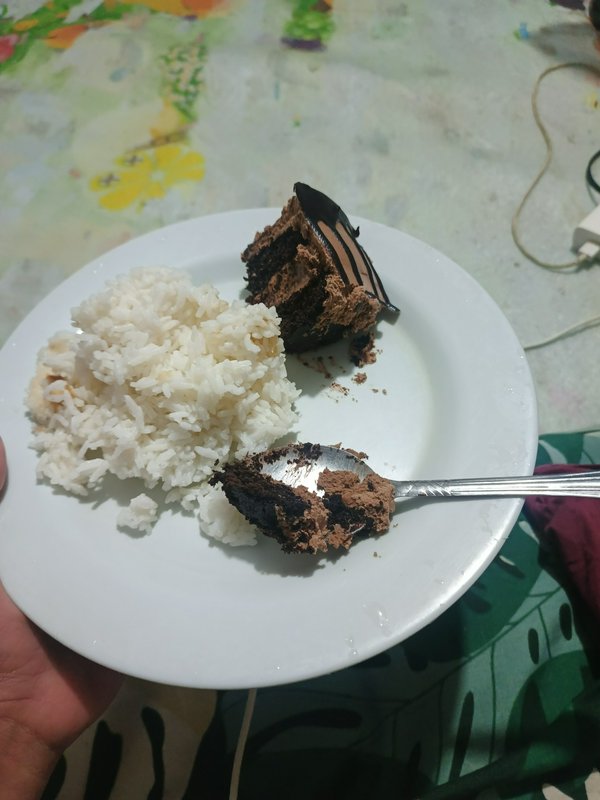 Hand holding white plate with fluffy steamed rice, partially eaten chocolate cake, and spoon with...