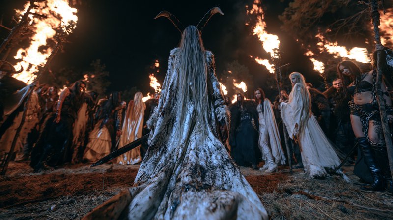 Back view of horned fantasy ritual leader standing before a gothic cult, flanked by blazing torch...