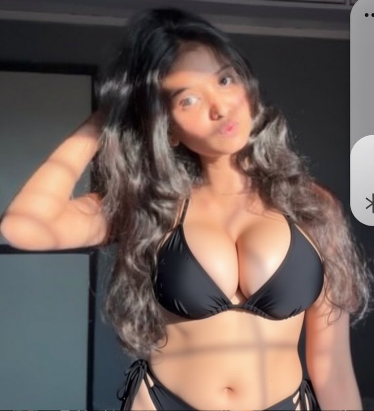 Young woman with long wavy gradient hair in black bikini, playful pout pose lit by warm indoor wi...