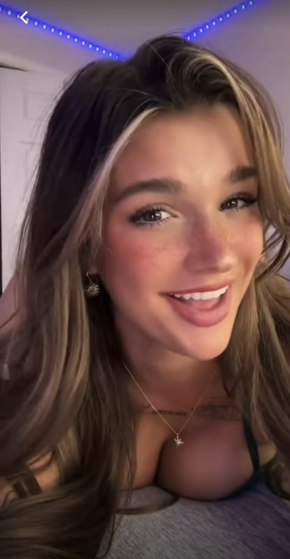 Close-up candid selfie of a smiling young woman with highlighted hair, star jewelry, in a bedroom...