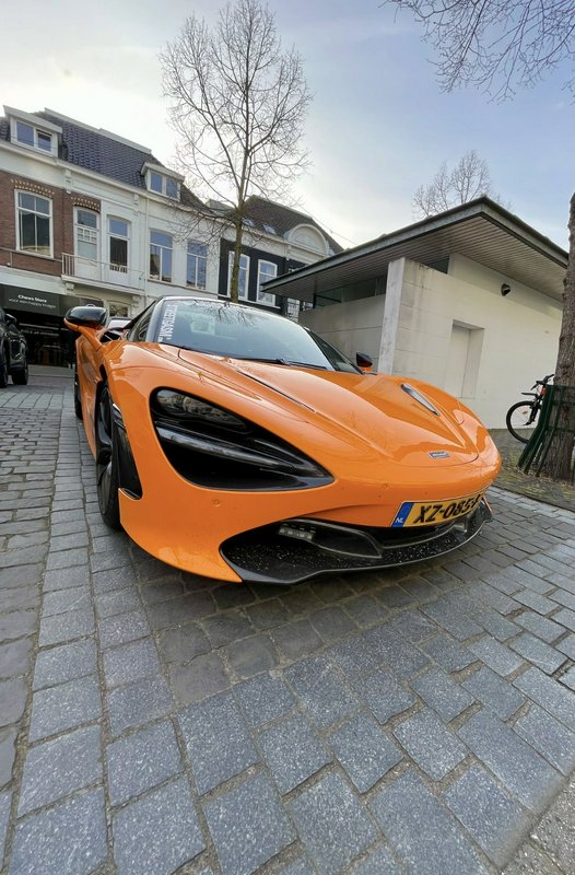 Low-angle view of a glossy orange McLaren supercar parked on a cobblestone European residential s...
