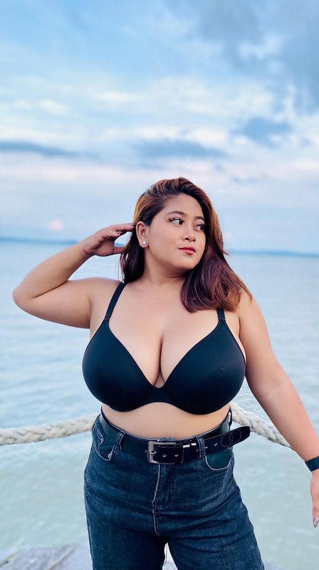 Curvy plus-size woman posing on a seaside dock, wearing a black plunge bra and jeans, with calm o...