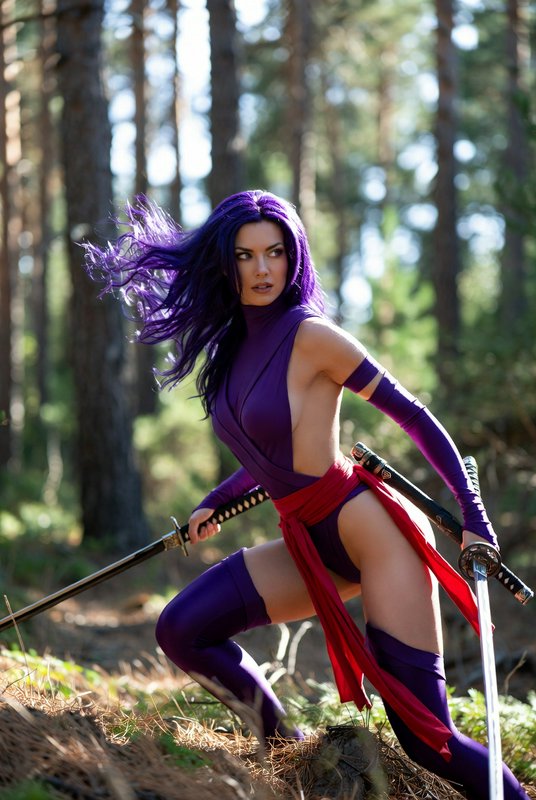 Female Psylocke cosplayer with flowing purple hair holds katanas in a combat stance in a sunlit pine forest